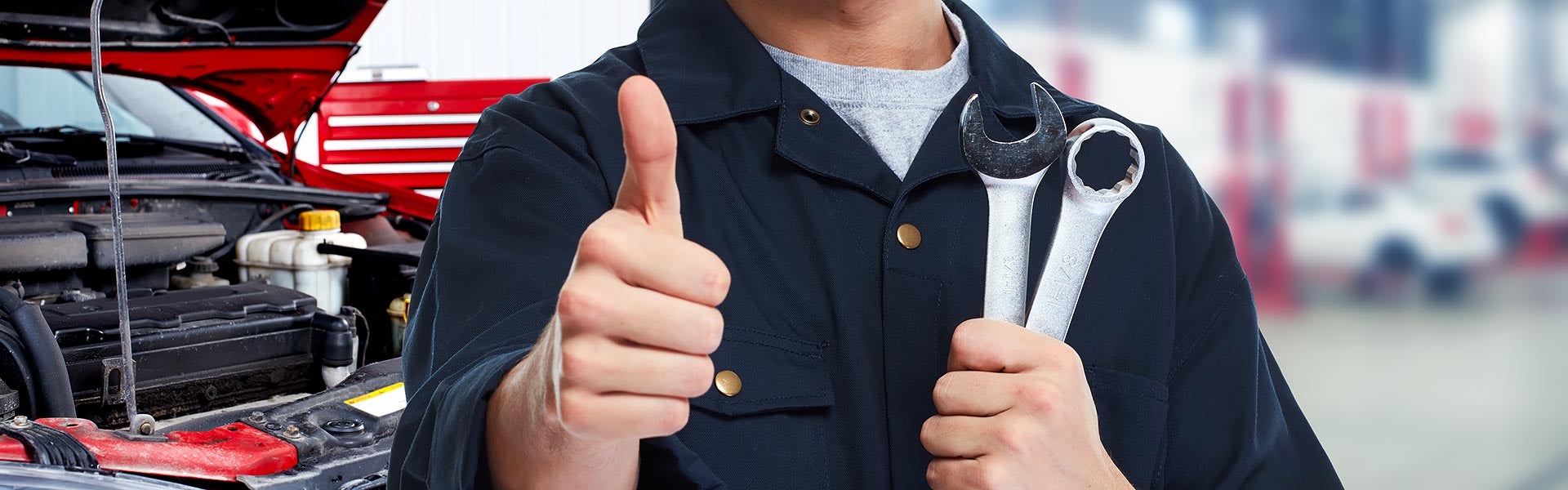 Mechanic holding wrenches and giving a thumbs-up next to an open car hood.